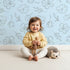 Lovely child standing in front of Whimsy Tots Petal Outline Bloom II Wallpaper in Sky Blue with flower, petal, bell flower, sprig, leaf, botanical in design. Lovely child standing in front of Whimsy Tots Petal Outline Bloom II Wallpaper in Sky Blue with flower, petal, bell flower, sprig, leaf, botanical in design.