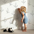 Lovely child standing in front of Whimsy Tots Whispering Leaflines Wallpaper in White with leaf, sprig, botanical, line art in design. Lovely child standing in front of Whimsy Tots Whispering Leaflines Wallpaper in White with leaf, sprig, botanical, line art in design.