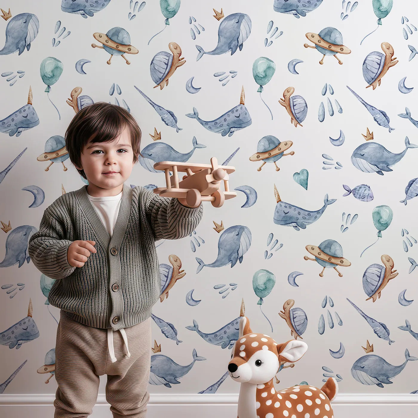 Lovely child standing in front of Whimsy Tots Galactic Ocean Friends Wallpaper in White with whale, turtle, narwhal, balloon, moon, UFO, droplet, crown in design.