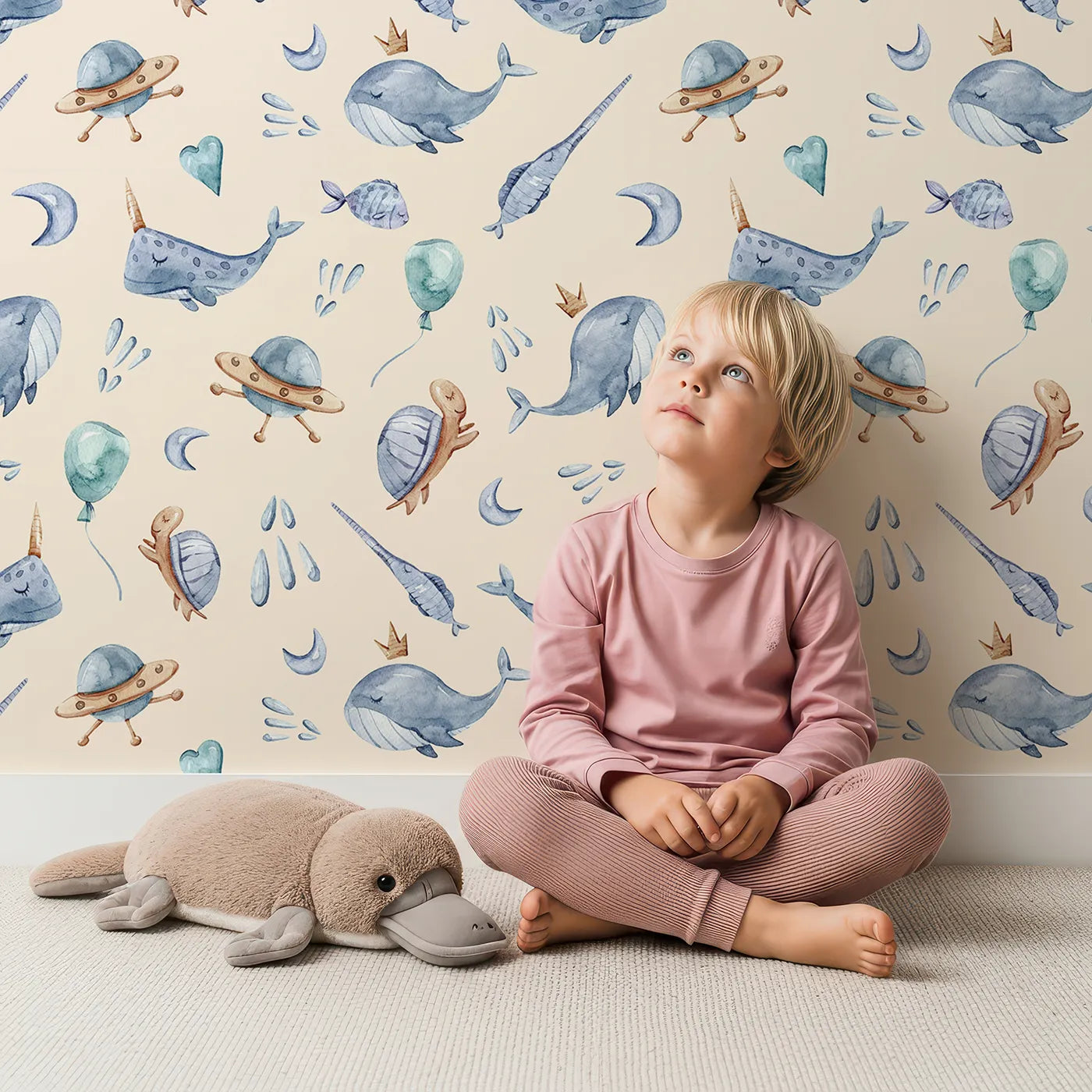 Lovely child standing in front of Whimsy Tots Galactic Ocean Friends Wallpaper in Sand with whale, turtle, narwhal, balloon, moon, UFO, droplet, crown in design.