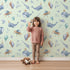 Lovely child standing in front of Whimsy Tots Galactic Ocean Friends Wallpaper in Mint Green with whale, turtle, narwhal, balloon, moon, UFO, droplet, crown in design. Lovely child standing in front of Whimsy Tots Galactic Ocean Friends Wallpaper in Mint Green with whale, turtle, narwhal, balloon, moon, UFO, droplet, crown in design.