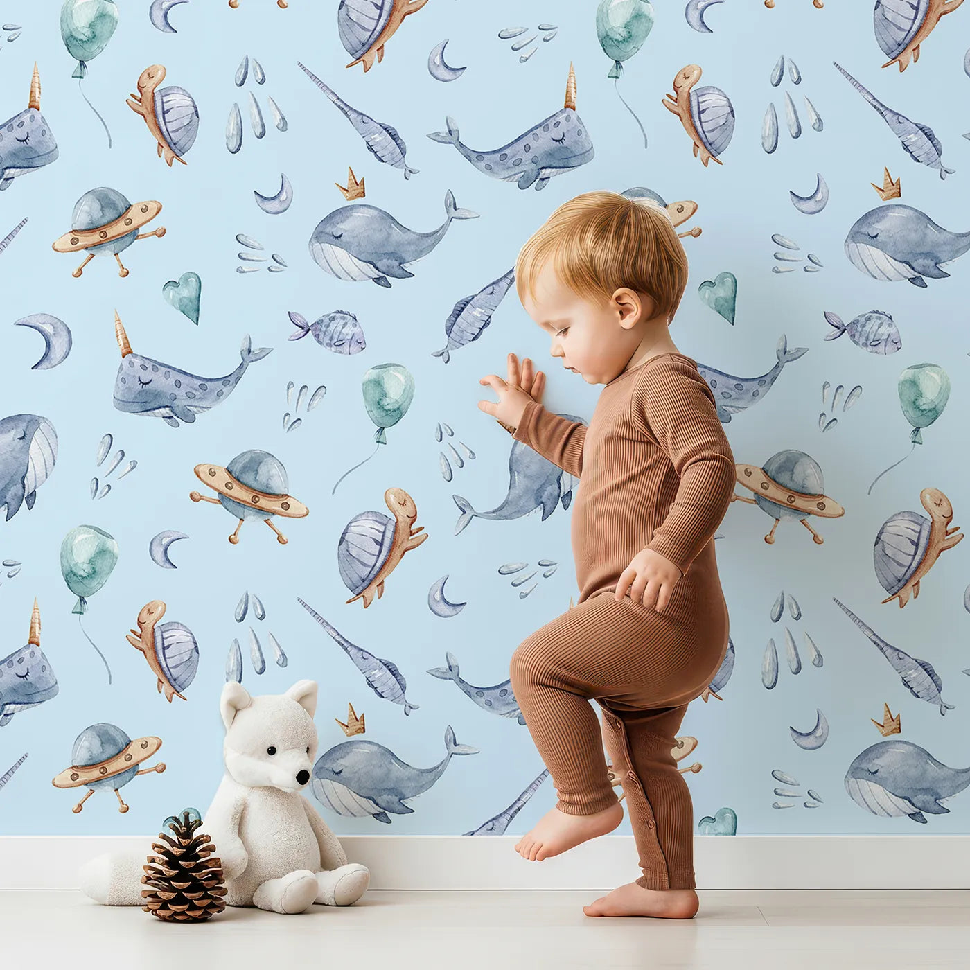 Lovely child standing in front of Whimsy Tots Galactic Ocean Friends Wallpaper in Blue with whale, turtle, narwhal, balloon, moon, UFO, droplet, crown in design.