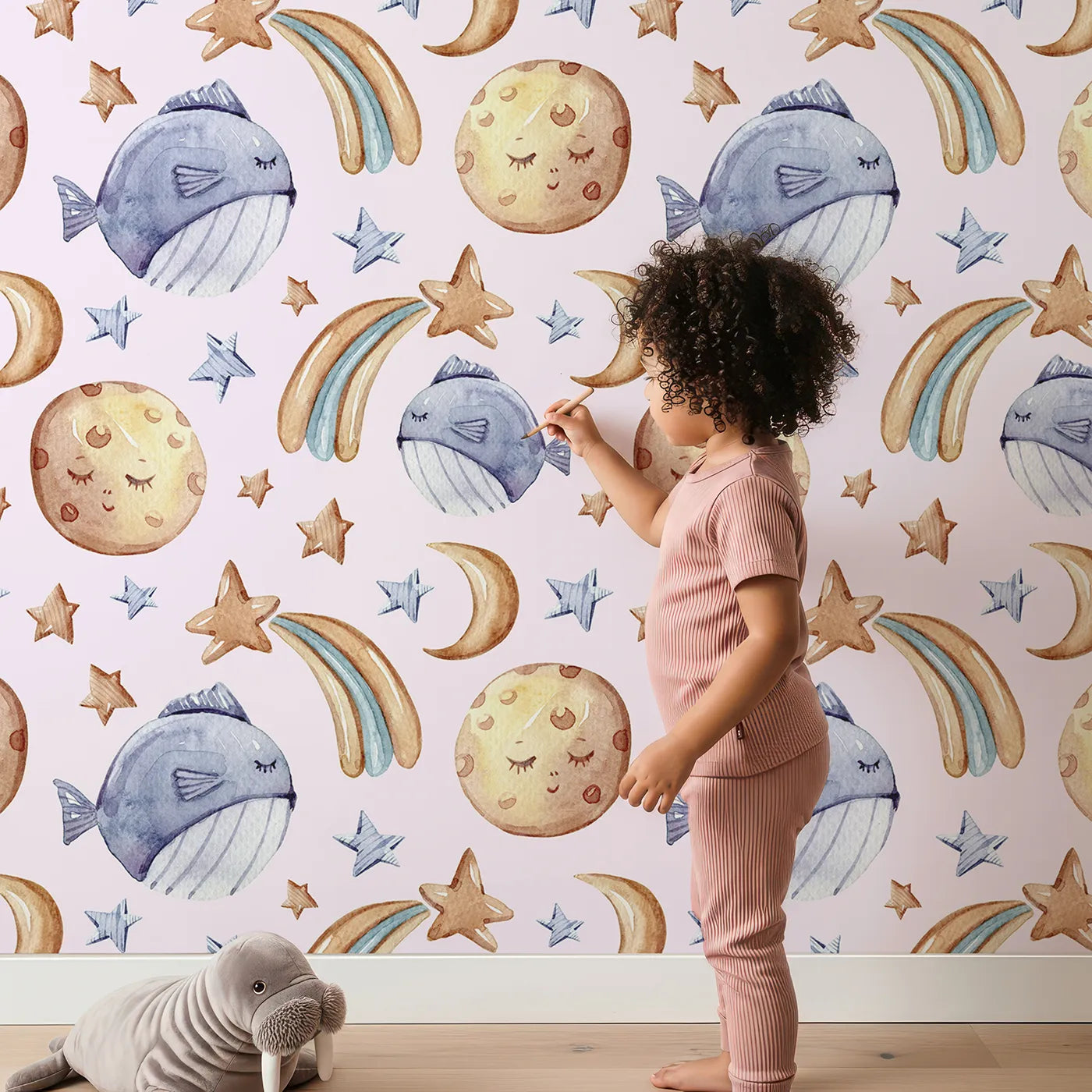 Lovely child standing in front of Whimsy Tots Sleepy Celestial Whale Wallpaper in Blush Pink with whale, moon, star, fish, comet in design.
