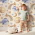 Lovely child standing in front of Whimsy Tots Sleepy Celestial Whale Wallpaper in Sand with whale, moon, star, fish, comet in design. Lovely child standing in front of Whimsy Tots Sleepy Celestial Whale Wallpaper in Sand with whale, moon, star, fish, comet in design.