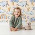 Lovely child standing in front of Whimsy Tots Sleepy Cloud Parade Wallpaper in Sand with cloud, moon, sheep, rainbow, star in design. Lovely child standing in front of Whimsy Tots Sleepy Cloud Parade Wallpaper in Sand with cloud, moon, sheep, rainbow, star in design.