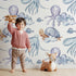 Lovely child standing in front of Whimsy Tots Ocean Cloud Cuddle Wallpaper in White with turtle, octopus, fish, cloud, squid in design. Lovely child standing in front of Whimsy Tots Ocean Cloud Cuddle Wallpaper in White with turtle, octopus, fish, cloud, squid in design.