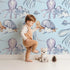 Lovely child standing in front of Whimsy Tots Ocean Cloud Cuddle Wallpaper in Sky Blue with turtle, octopus, fish, cloud, squid in design. Lovely child standing in front of Whimsy Tots Ocean Cloud Cuddle Wallpaper in Sky Blue with turtle, octopus, fish, cloud, squid in design.