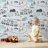 Lovely child standing in front of Whimsy Tots Animal Train Adventure Wallpaper in White with train, lion, elephant, tiger, rainbow, tree, mountain in design. Lovely child standing in front of Whimsy Tots Animal Train Adventure Wallpaper in White with train, lion, elephant, tiger, rainbow, tree, mountain in design.
