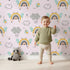 Lovely child standing in front of Whimsy Tots Bee Rainbow Dreams Wallpaper in Blush Pink with bee, rainbow, star, cloud in design. Lovely child standing in front of Whimsy Tots Bee Rainbow Dreams Wallpaper in Blush Pink with bee, rainbow, star, cloud in design.