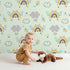 Lovely child standing in front of Whimsy Tots Bee Rainbow Dreams Wallpaper in Mint Green with bee, rainbow, star, cloud in design. Lovely child standing in front of Whimsy Tots Bee Rainbow Dreams Wallpaper in Mint Green with bee, rainbow, star, cloud in design.