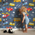 Lovely child standing in front of Whimsy Tots Vroom Vroom Transport Parade Wallpaper in Navy Blue with bus, train, plane, car, helicopter, cloud in design. Lovely child standing in front of Whimsy Tots Vroom Vroom Transport Parade Wallpaper in Navy Blue with bus, train, plane, car, helicopter, cloud in design.