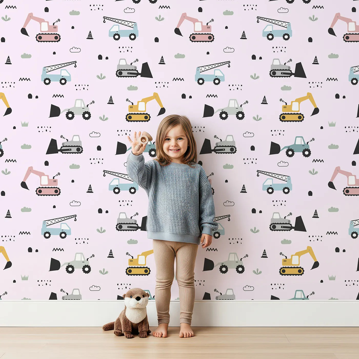 Lovely child standing in front of Whimsy Tots Digger Parade Wallpaper in Pink with crane, digger, loader, truck, cone in design.