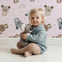 Lovely child standing in front of Whimsy Tots Koala Cuddle Parade Wallpaper in Pink with koala, rainbow, heart, raindrop in design. Lovely child standing in front of Whimsy Tots Koala Cuddle Parade Wallpaper in Pink with koala, rainbow, heart, raindrop in design.