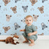 Lovely child standing in front of Whimsy Tots Koala Cuddle Parade Wallpaper in Blue with koala, rainbow, heart, raindrop in design. Lovely child standing in front of Whimsy Tots Koala Cuddle Parade Wallpaper in Blue with koala, rainbow, heart, raindrop in design.