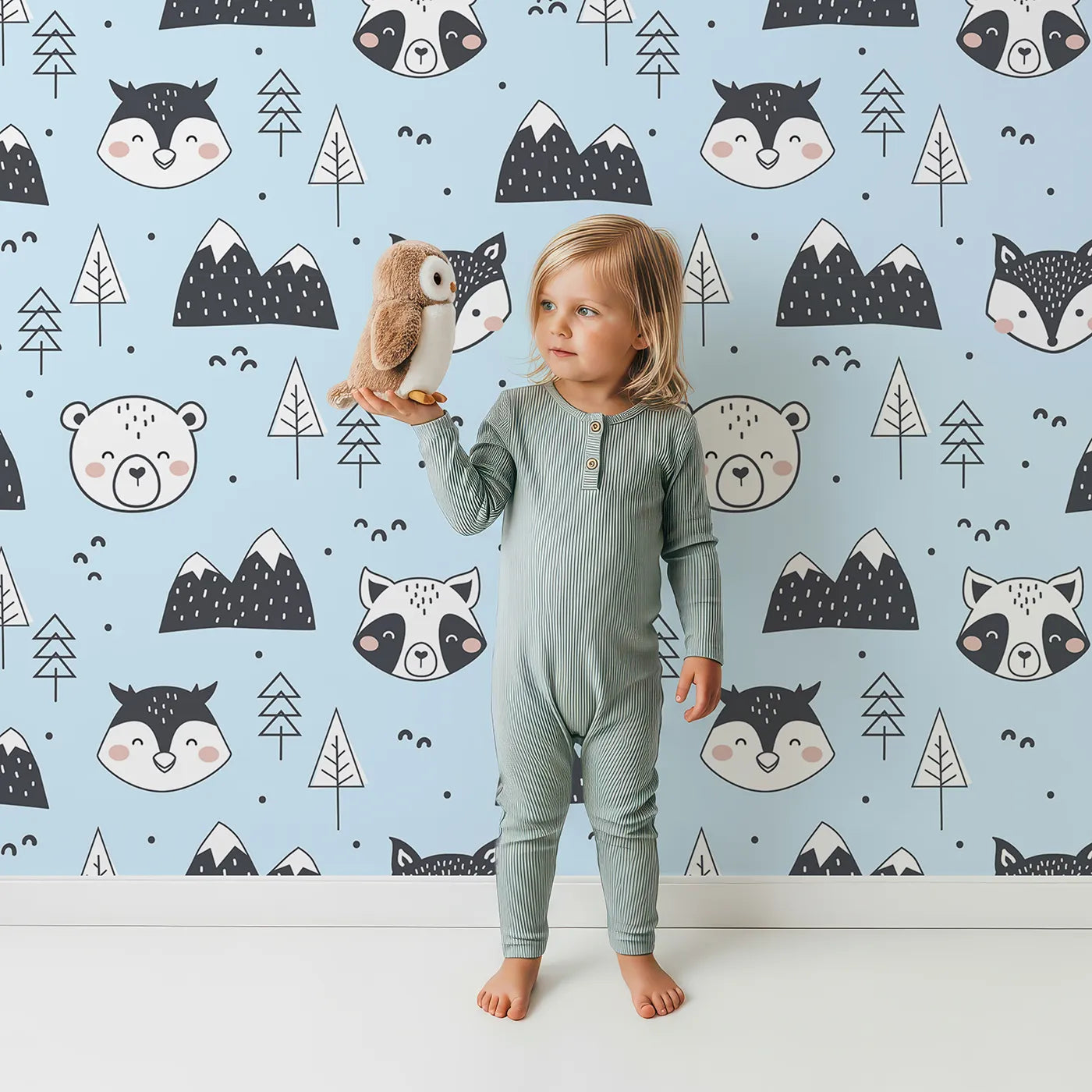 Lovely child standing in front of Whimsy Tots Woodland Peekaboo Friends Wallpaper in Blue with bear, owl, fox, raccoon, mountain, tree in design.