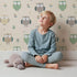 Lovely child standing in front of Whimsy Tots Gentle Owl Garden Wallpaper in Warm Cream with owl, flower, leaf in design. Lovely child standing in front of Whimsy Tots Gentle Owl Garden Wallpaper in Warm Cream with owl, flower, leaf in design.