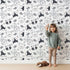 Lovely child standing in front of Whimsy Tots Woodland Scribble Friends Wallpaper in Monochrome with bear, bunny, fox, raccoon, flower, leaf in design. Lovely child standing in front of Whimsy Tots Woodland Scribble Friends Wallpaper in Monochrome with bear, bunny, fox, raccoon, flower, leaf in design.