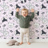 Lovely child standing in front of Whimsy Tots Woodland Scribble Friends Wallpaper in Blush Pink with bear, bunny, fox, raccoon, flower, leaf in design. Lovely child standing in front of Whimsy Tots Woodland Scribble Friends Wallpaper in Blush Pink with bear, bunny, fox, raccoon, flower, leaf in design.