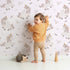 Lovely child standing in front of Whimsy Tots Feathered DaydreamโฏUnicorn Wallpaper in Blushย Pink with unicorn, feather, moon, star, leaf in design. Lovely child standing in front of Whimsy Tots Feathered DaydreamโฏUnicorn Wallpaper in Blushย Pink with unicorn, feather, moon, star, leaf in design.