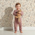 Lovely child standing in front of Whimsy Tots Whispering Ferns Wallpaper in Sandstone with fern, leaf, branch, foliage in design. Lovely child standing in front of Whimsy Tots Whispering Ferns Wallpaper in Sandstone with fern, leaf, branch, foliage in design.
