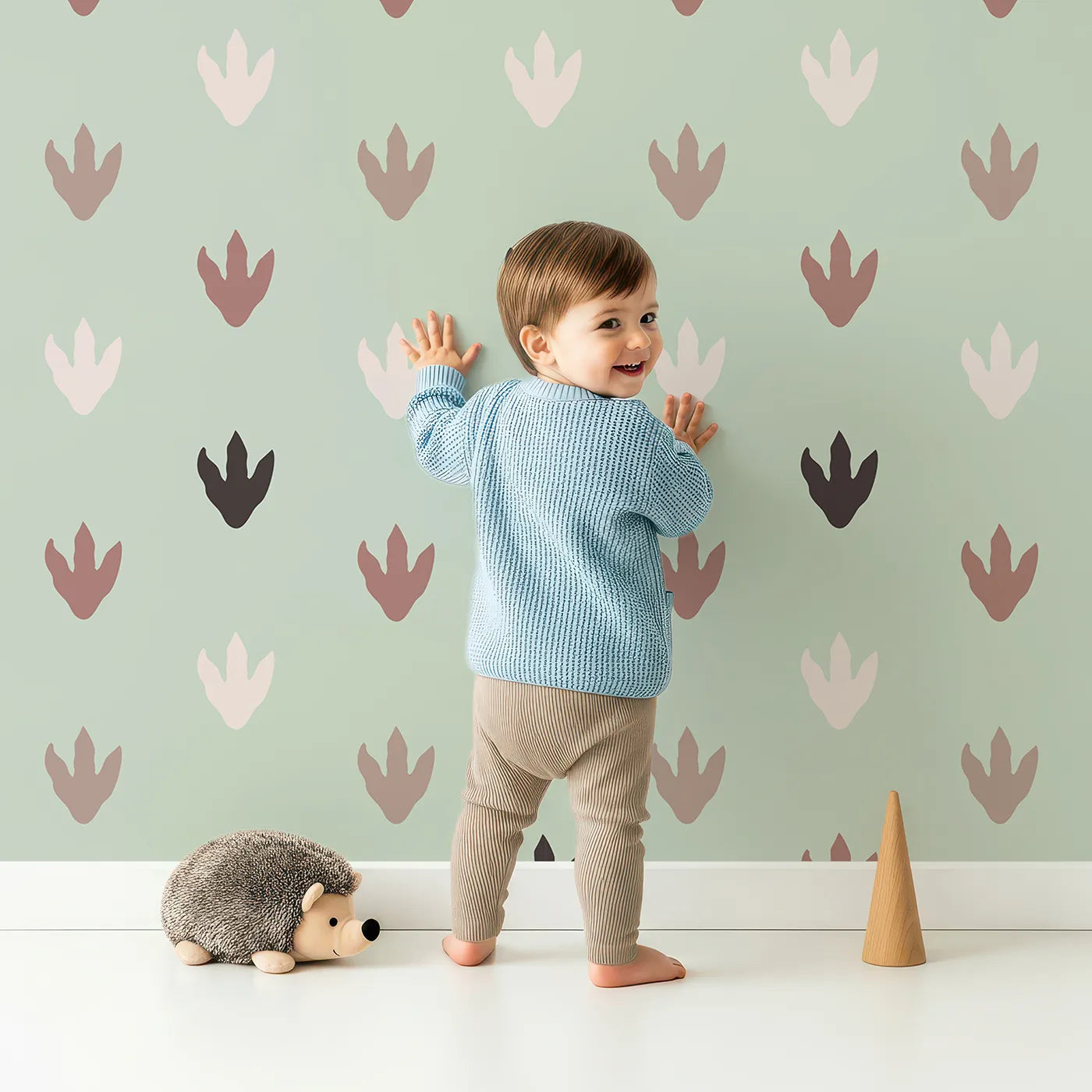 Lovely child standing in front of Whimsy Tots Dino Tracks Wallpaper in Sage Green with footprint, dinosaur, track, print in design.
