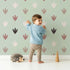 Lovely child standing in front of Whimsy Tots Dino Tracks Wallpaper in Sage Green with footprint, dinosaur, track, print in design. Lovely child standing in front of Whimsy Tots Dino Tracks Wallpaper in Sage Green with footprint, dinosaur, track, print in design.