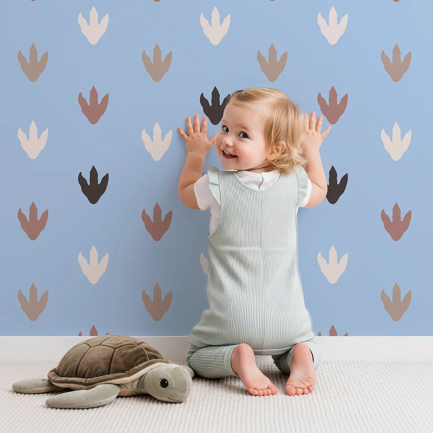 Lovely child standing in front of Whimsy Tots Dino Tracks Wallpaper in Sky Blue with footprint, dinosaur, track, print in design.