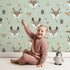 Lovely child standing in front of Whimsy Tots Forest Forager Deer Wallpaper in Sage Green with deer, pinecone, tree, snowflake, berry in design. Lovely child standing in front of Whimsy Tots Forest Forager Deer Wallpaper in Sage Green with deer, pinecone, tree, snowflake, berry in design.