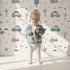 Lovely child standing in front of Whimsy Tots Dino Drivers Parade Wallpaper in White with dinosaur, car, tree, rainbow, mountain in design. Lovely child standing in front of Whimsy Tots Dino Drivers Parade Wallpaper in White with dinosaur, car, tree, rainbow, mountain in design.