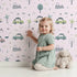 Lovely child standing in front of Whimsy Tots Dino Drivers Parade Wallpaper in Blush Pink with dinosaur, car, tree, rainbow, mountain in design. Lovely child standing in front of Whimsy Tots Dino Drivers Parade Wallpaper in Blush Pink with dinosaur, car, tree, rainbow, mountain in design.