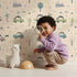 Lovely child standing in front of Whimsy Tots Dino Drivers Parade Wallpaper in Cream with dinosaur, car, tree, rainbow, mountain in design. Lovely child standing in front of Whimsy Tots Dino Drivers Parade Wallpaper in Cream with dinosaur, car, tree, rainbow, mountain in design.