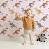 Lovely child standing in front of Whimsy Tots Gentle Dino Trek Wallpaper in Blush Pink with dinosaur, footprint, plant in design. Lovely child standing in front of Whimsy Tots Gentle Dino Trek Wallpaper in Blush Pink with dinosaur, footprint, plant in design.