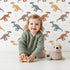 Lovely child standing in front of Whimsy Tots Gentle Dino Trek Wallpaper in White with dinosaur, footprint, plant in design. Lovely child standing in front of Whimsy Tots Gentle Dino Trek Wallpaper in White with dinosaur, footprint, plant in design.