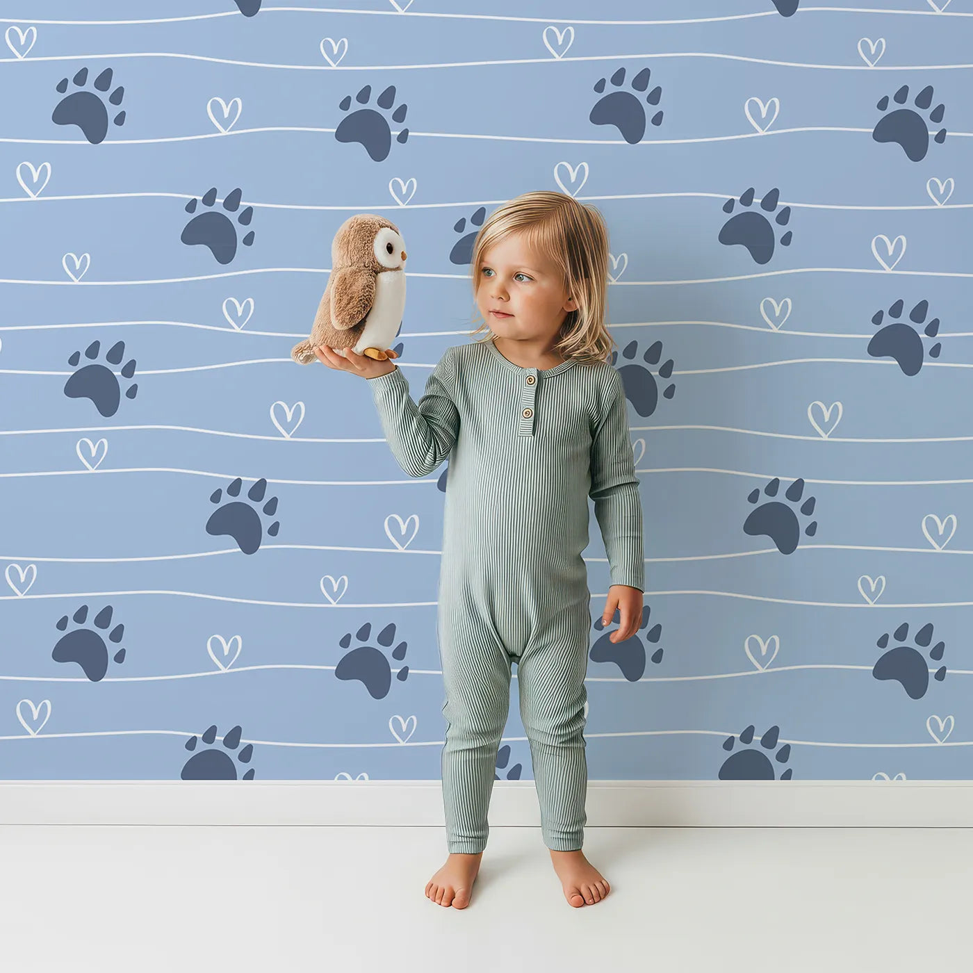 Lovely child standing in front of Whimsy Tots Bear Hug Trail Wallpaper in Sky Blue with bear, paw print, heart, line in design.