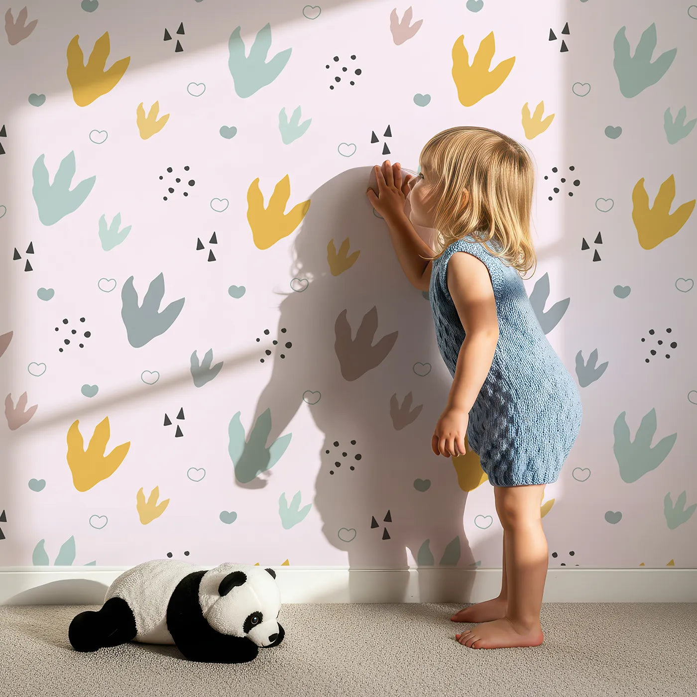 Lovely child standing in front of Whimsy Tots Dino Stomp Parade Wallpaper in Blush Pink with dinosaur, footprint, heart, dot, triangle in design.
