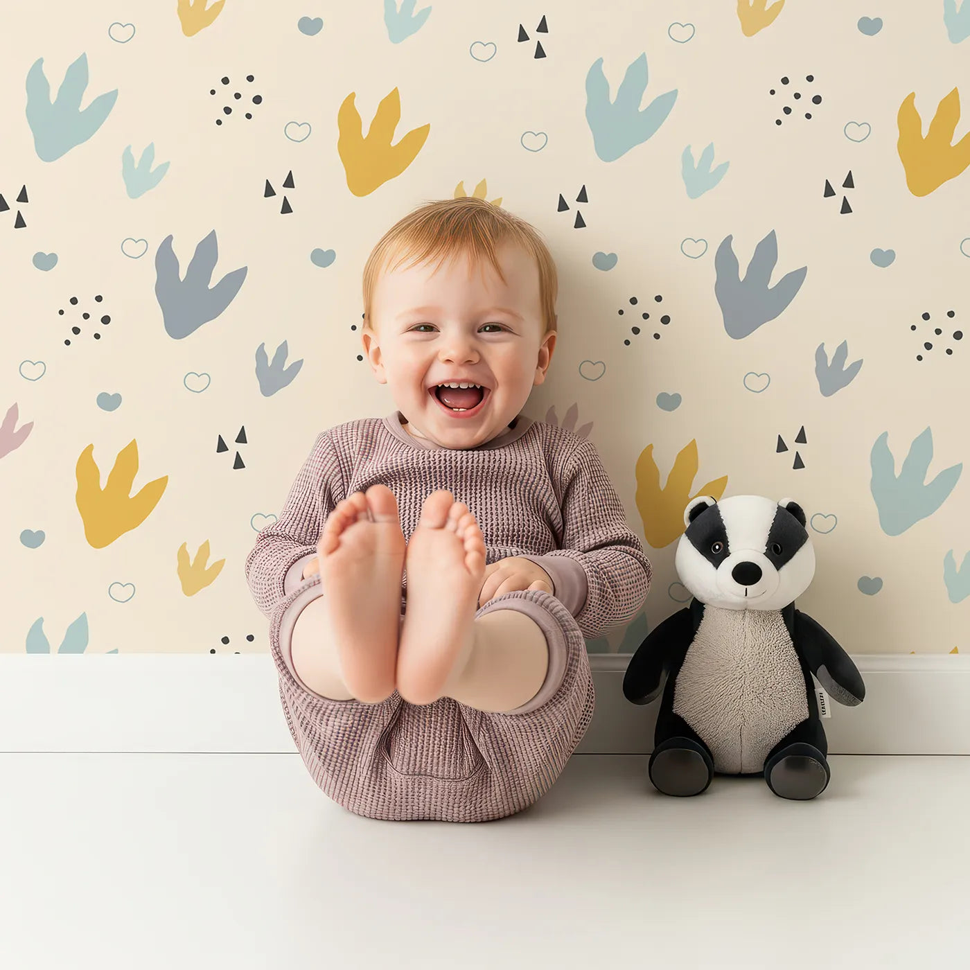 Lovely child standing in front of Whimsy Tots Dino Stomp Parade Wallpaper in Soft Sandstone with dinosaur, footprint, dot, triangle, heart in design.