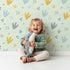 Lovely child standing in front of Whimsy Tots Dino Stomp Parade Wallpaper in Mint Green with dinosaur, footprint, dot, triangle, heart in design. Lovely child standing in front of Whimsy Tots Dino Stomp Parade Wallpaper in Mint Green with dinosaur, footprint, dot, triangle, heart in design.