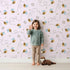 Lovely child standing in front of Whimsy Tots Busy Bee Trail Wallpaper in Blush Pink with bee, flower, leaf, heart, trail in design. Lovely child standing in front of Whimsy Tots Busy Bee Trail Wallpaper in Blush Pink with bee, flower, leaf, heart, trail in design.
