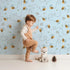 Lovely child standing in front of Whimsy Tots Busy Bee Trail Wallpaper in Sky Blue with bee, flower, leaf, heart, trail in design. Lovely child standing in front of Whimsy Tots Busy Bee Trail Wallpaper in Sky Blue with bee, flower, leaf, heart, trail in design.