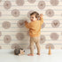Lovely child standing in front of Whimsy Tots Boho Rhythm Doodle Wallpaper in Soft Beige with rainbow, heart, star, stripe, doodle in design. Lovely child standing in front of Whimsy Tots Boho Rhythm Doodle Wallpaper in Soft Beige with rainbow, heart, star, stripe, doodle in design.