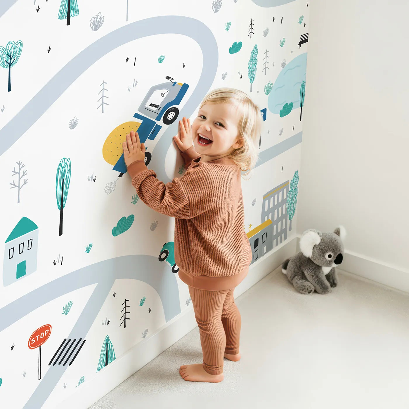 Child in front of Whimsy Tots Busy Town Roads Wall Mural with car, road, town, city, house, tree, truck, bus in design.