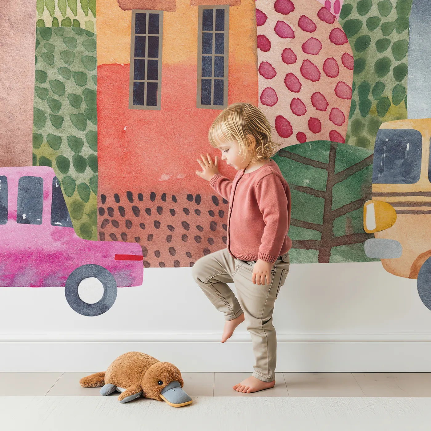Child in front of Whimsy Tots Watercolor Town Street Wall Mural with house, town, car, bus, watercolor, street, city, pink in design.