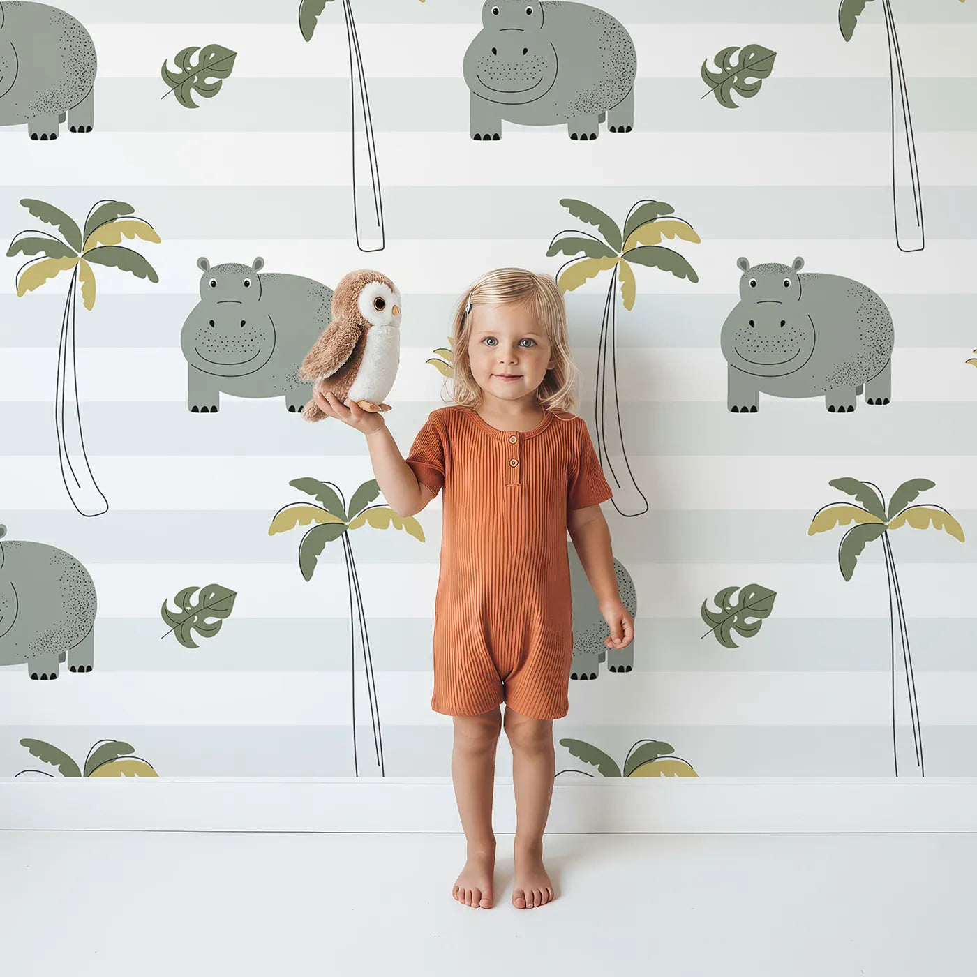 Lovely child standing in front of Whimsy Tots Striped Hippo Jungle Wallpaper with hippo, palm tree, tropical, jungle, stripe in design.