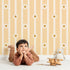 Lovely child standing in front of Whimsy Tots Daisy Lane Stripe Wallpaper in Yellow with daisy, flower, stripe in design.