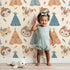 Lovely child standing in front of Whimsy Tots Tribal Pals Wallpaper in Cream with owl, sloth, deer, tiger, tent, feather in design.