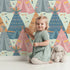 Lovely child standing in front of Whimsy Tots Teepee Parade Wallpaper in Mint Green with teepee, arrow, triangle, tent in design.