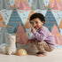 Lovely child standing in front of Whimsy Tots Teepee Parade Wallpaper in Blue with teepee, arrow, triangle, tent in design.