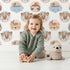 Lovely child standing in front of Whimsy Tots Woodland Royalty Parade Wallpaper in White with bear, deer, elephant, tiger, feather, wreath, crown in design.