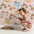 Lovely child standing in front of Whimsy Tots Wild Whimsy Tribe Wallpaper in Pink with bear, deer, elephant, tiger, owl, sloth, arrow, feather, star in design.