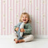 Lovely child standing in front of Whimsy Tots Milky Morning Stripes Wallpaper in Pink with milk, stripe, bottle in design.